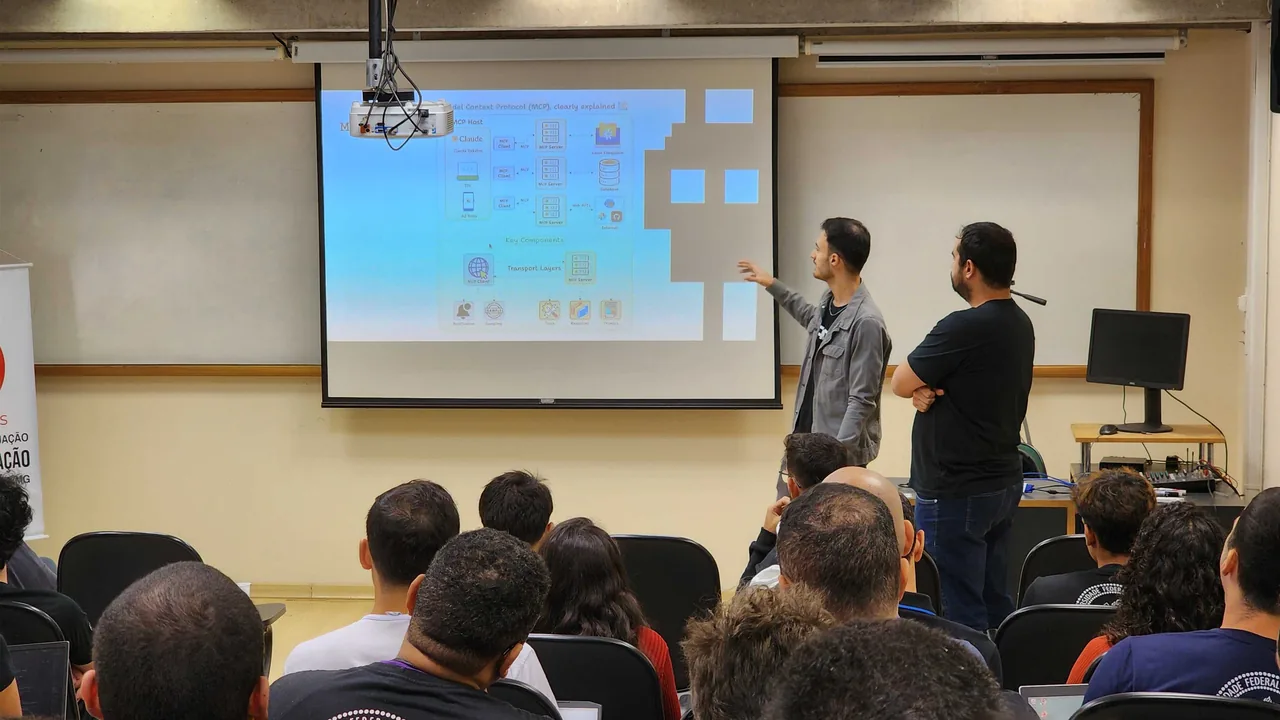Two instructors at a projected slide explaining the Model Context Protocol, one pointing at the diagram, with a classroom of students watching